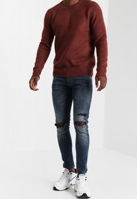 Maglione lavorato a maglia di colore ruggine, peso medio, collo tondo. Jeans blu strappati con buchi sulle ginocchia. Scarpe da ginnastica bianche con dettagli neri.