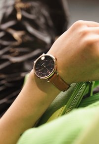 Montre Fossil ronde avec un cadran noir, un boîtier en or rose et un bracelet en cuir marron, présentée sur une main à côté d'un sac à main vert.