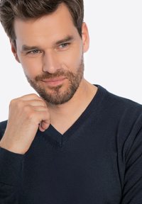 Granatowy sweter w serek z miękkiego dzianiny, z prążkowanymi mankietami i dekoltem. Modelka przedstawiona w zamyślonej pozie, z ręką przy brodzie.