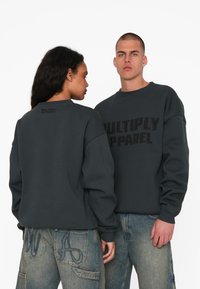 Sweatshirt oversize gris foncé avec "MULTIPLY APPAREL" imprimé en noir. Présente des poignets côtelés et un col ras du cou, porté avec un pantalon en denim clair.