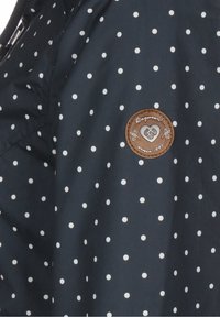 Mörkblå jacka med vita prickar, med en rund brunt lädermärke med präglade detaljer, slät textur och dragkedja.