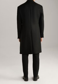 Cappotto di lana nera con un design sartoriale, silhouette lunga e apertura singola. Abbinato a pantaloni neri e scarpe lucide.