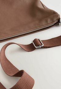 Braune Ledertasche mit einem silbernen Reißverschluss, ausgestattet mit einem passenden braunen Stoffgurt und Metalldetails. Die Textur ist glatt mit sichtbaren Nähten.