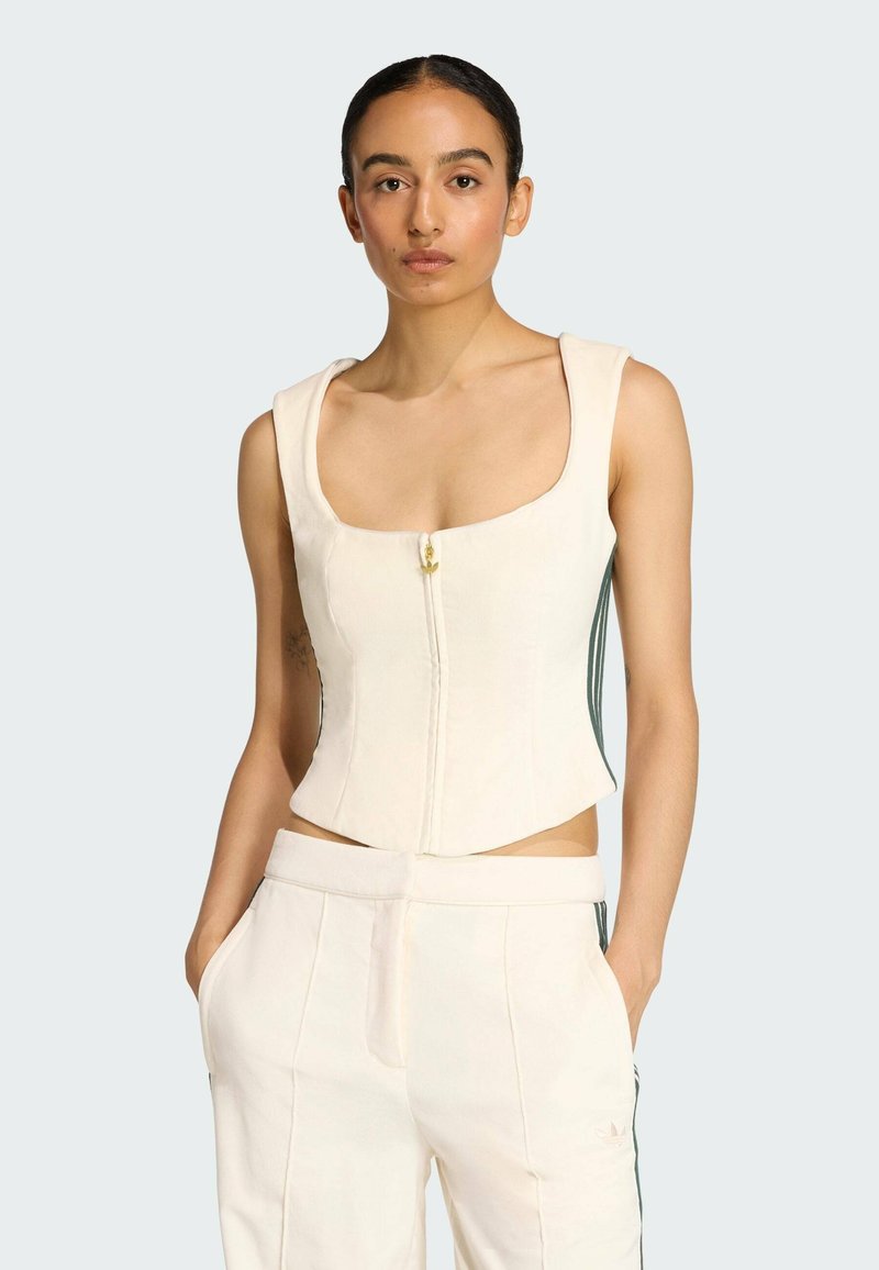 Crème-kleurige cropped top met vierkante halslijn, rits aan de voorkant en een gouden knoopdetail. Draagt met bijpassende lichte broek met zijstrepen.