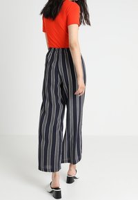 Kvinna som bär en röd kortärmad topp, marinblå vida byxor med vita vertikala ränder och vita klackmules, går bort.