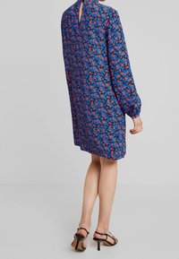 Robe fleurie avec un fond bleu orné de fleurs rouges et bleues, manches longues, et une découpe en forme de trou de serrure au dos. Longueur au-dessus du genou.