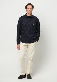 Camicia a maniche lunghe blu navy con due tasche a patta e bottoni a pressione argento, abbinata a pantaloni color crema e scarpe nere.