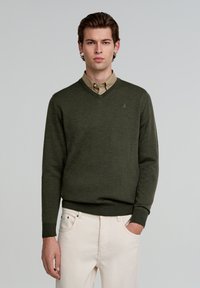 Pulover verde cu decolteu în V, cu un finisaj texturat, purtat peste o cămașă deschisă la culoare, beige, cu guler. Puloverul are manșete ribbate și un detaliu cu logo.