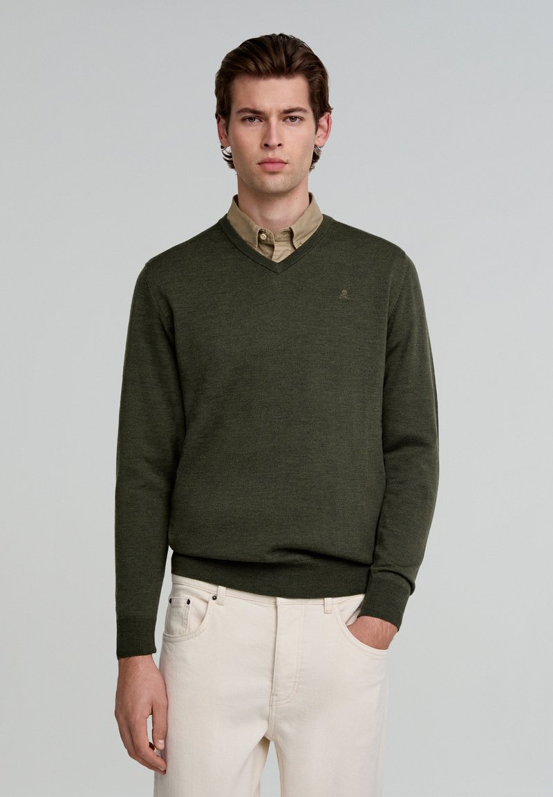 Pulover verde cu decolteu în V, cu un finisaj texturat, purtat peste o cămașă deschisă la culoare, beige, cu guler. Puloverul are manșete ribbate și un detaliu cu logo.