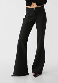 Černé kalhoty s vysokým pasem a rozšiřujícími se nohavicemi vyrobené z hladké látky, s předním zipem pro zapínání a přizpůsobeným střihem, který prodlužuje nohy.