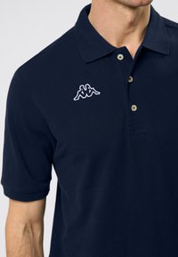 Tamsiai mėlyna polo marškinėlių su tekstūruotu audiniu. Su trimis mygtukais priekyje ir išsiuvinėtu logotipu kairėje krūtinės pusėje. Trumpomis rankovėmis.