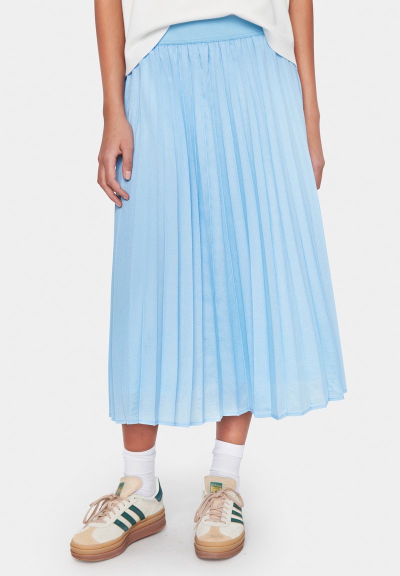 Vaaleansininen laskostettu hame, joka on valmistettu tasaisesta kankaasta. Hameessa on vyötäröosa, ja se ulottuu polven keskikohtaan. Yhdistettynä valkoisiin sukkiin ja rentoihin tennareihin.