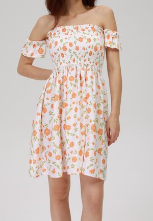 Kvinde iført en hvid skulderafslørende kjole med orange blomsterprint og flæseærmer, stående foran en ensfarvet baggrund.