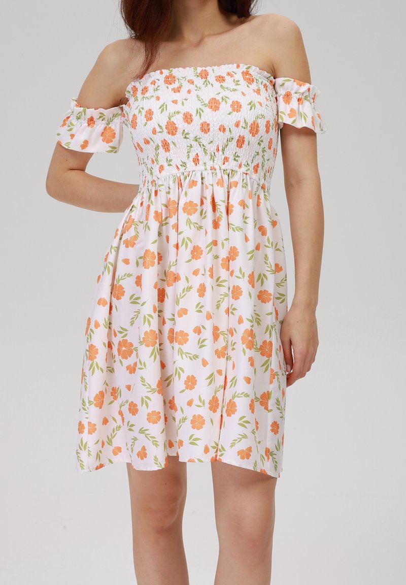 Femme portant une robe blanche épaules dénudées avec un imprimé floral orange et des manches à volants, debout devant un fond uni.