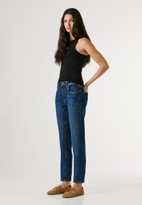 Jauna sieviete ar gariem matiem, valkājot melnu krekliņu bez piedurknēm, zilas džinsas un brūnas loafers kurpes, stāvot ar vienu roku kabatā.