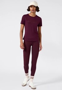 Bordeauxkleurig sport-T-shirt met korte mouwen en logo, gecombineerd met bijpassende leggings. Witte sneakers met bruine accenten en gestreepte sokken.