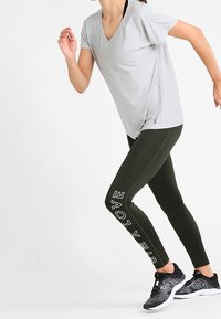 T-shirt grigio chiaro con scollo a v, leggings verde oliva con testo bianco "LOVE", sneaker nere con dettagli testurizzati e una posa di corsa dinamica.