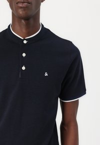 Marineblauwe poloshirt met korte mouwen en een Mandarin-kraag, witte knoopaccenten en een subtiel logo op de borst. Voorzien van contrasterende witte afwerking.