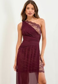 Frau in einem burgunderfarbenen, einseitigen Kleid mit Falten und Spitzenbesatz, die vor einem schlichten, hellen Hintergrund steht.