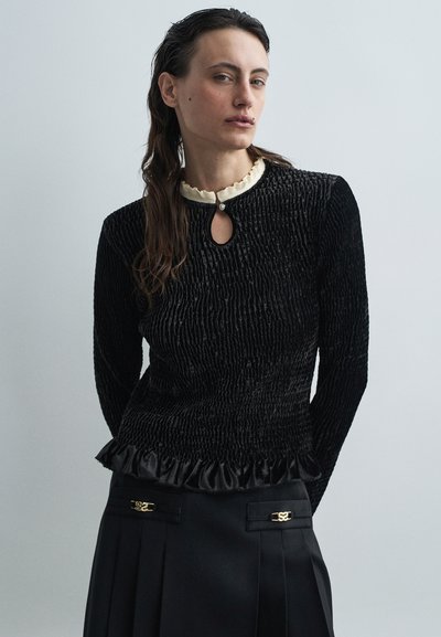 Haut noir texturé avec un col à découpe, col en crêpe crème à volants et ourlet volant en satin, associé à une jupe plissée noire avec des accents dorés.