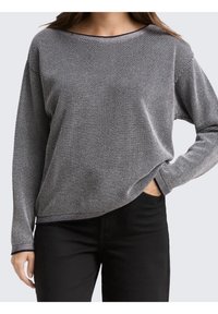 Femme portant un pull manches longues gris et noir à motifs avec un pantalon noir, main dans la poche, debout devant un fond uni.