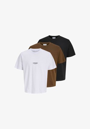 Un jeune homme se tient debout, portant un t-shirt blanc JJ Studios, un pantalon beige et des baskets marron sur un fond clair uni.