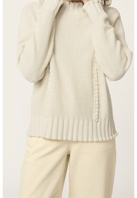 Eine Person trägt einen cremefarbenen Strickpullover mit geripptem Saum und passenden cremefarbenen Hosen. Die Hände befinden sich in der Nähe des Gesichts, vor einem schlichten hellen Hintergrund.
