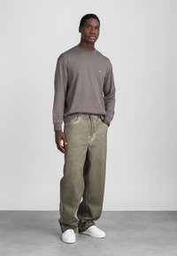 Hallikas pika varrukaga särk väikese logoga, koos vabade oliivroheliste pükstega ja valgete tossudega, neutraalses taustas.