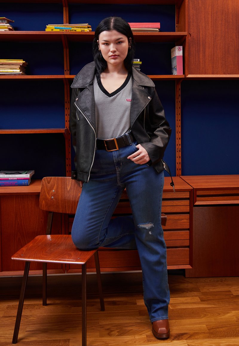 Giacca in ecopelle nera da moto, t-shirt a righe con scollo a V, jeans blu a vita alta con strappi, cintura nera con fibbia dorata, scarpe marroni con tacco a blocco.