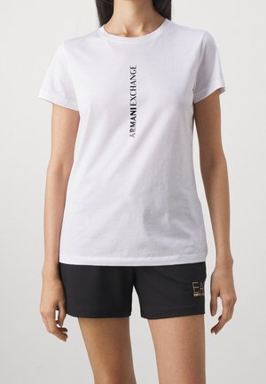 Kobieta nosząca białą koszulkę z krótkim rękawem z pionowym tekstem "Armani Exchange" oraz czarne szorty z logo "EA7 Emporio Armani" na lewej nogawce.