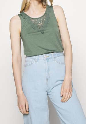 Oliven grøn tanktop med et blonde-design omkring udskæringen, parret med lyseblå højtaljede denimjeans.