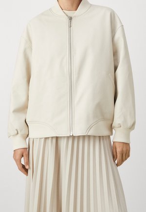 Personne portant une veste bomber crème à fermeture éclair sur une jupe plissée beige, debout devant un fond blanc uni.