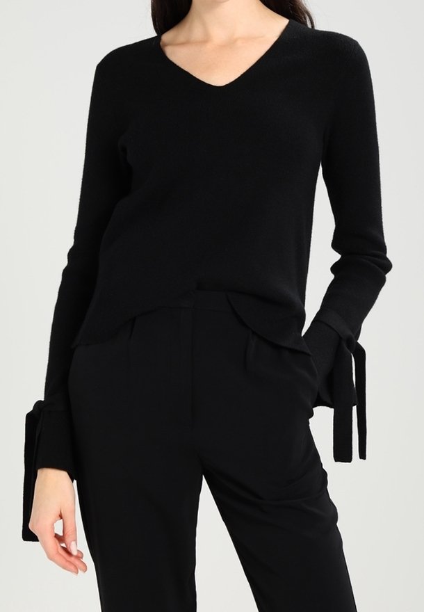 Black knitted sweater with a V-neck, long sleeves featuring tie accents at the cuffs, and a relaxed fit. Paired with black trousers.