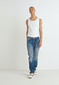 Top in cotone bianco a coste, jeans blu strappati con tasche e sneakers beige. Il modello è in piedi contro uno sfondo chiaro e semplice.