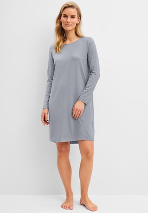 Vrouw met blond haar die een losse, lange mouwen, grijze jurk tot op de knie draagt en blootsvoets staat tegen een eenvoudige lichte achtergrond.
