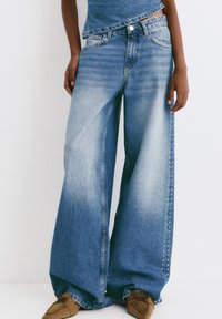 Blugi cu croială largă din denim albastru, cu o nuanță decolorată, având studs metallici pe cusăturile exterioare, asortați cu un top casual.