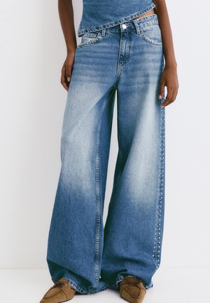 Weite blaue Jeans aus Denim mit einem ausgewaschenen Finish, versehen mit metallischen Nieten entlang der äußeren Nähte, kombiniert mit einem legeren Oberteil.