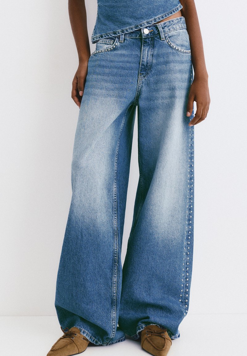 Blugi cu croială largă din denim albastru, cu o nuanță decolorată, având studs metallici pe cusăturile exterioare, asortați cu un top casual.