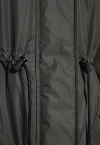 Tmavě zelená venkovní bunda s hladkou, mírně lesklou texturou. Na přední straně má stahovací šňůrky s černými posuvnými pojistkami.