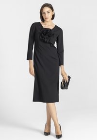 Czarna sukienka z długimi rękawami, dopasowany gorset z dużym falbanianym detalem, projekt o długości do kolan, w zestawie z czarną kopertówką i szpilkami.