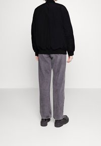 Blouson aviateur noir avec poignets et ourlet côtelés, associé à un pantalon en denim gris ample et à des chaussures à plateforme noires. Design simple et texturé.