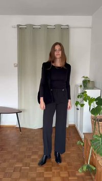 Blusa preta justa de manga longa e calças largas de cintura alta, combinadas com botas pretas de cano curto. Ambiente interior com uma cortina leve e plantas.