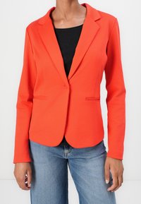 Blazer ajustado rojo con solapas, cierre de un solo botón y dos bolsillos frontales, llevado sobre una blusa negra, combinado con jeans azules.