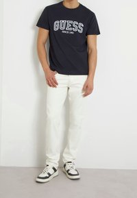 Camiseta de algodón azul marino con el texto "GUESS SINCE 1981"; jeans blancos de mezclilla; zapatillas negras y blancas; de pie contra un fondo liso.