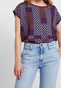 Top de manga corta con un diseño geométrico en púrpura oscuro, naranja y rosa, combinado con unos pantalones vaqueros de tiro alto en azul claro.