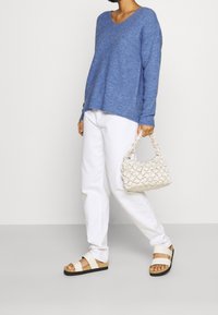 Personne portant un pull bleu et un pantalon blanc, tenant un sac à main crème, se tenant sur un fond neutre avec des sandales décontractées.