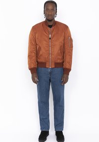 Bronzen bomberjack met ribgebreide manchetten en zoom, ritszakken, gedragen over een zwart shirt, gecombineerd met blauwe spijkerbroeken en zwarte schoenen.