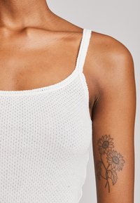 Top a canottiera bianco a coste con bretelle sottili, realizzato in tessuto texturizzato e con un bordo scallop. Un tatuaggio a forma di girasole è visibile sul braccio.