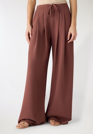 Donna che indossa pantaloni ampi a vita alta color ruggine con dettaglio annodato sul davanti e sandali beige, in piedi davanti a uno sfondo semplice.
