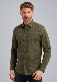 Chemise à manches longues verte à motif, avec un col à boutons, présentant une surface texturée et des boutons noirs, portée sur un jean noir.